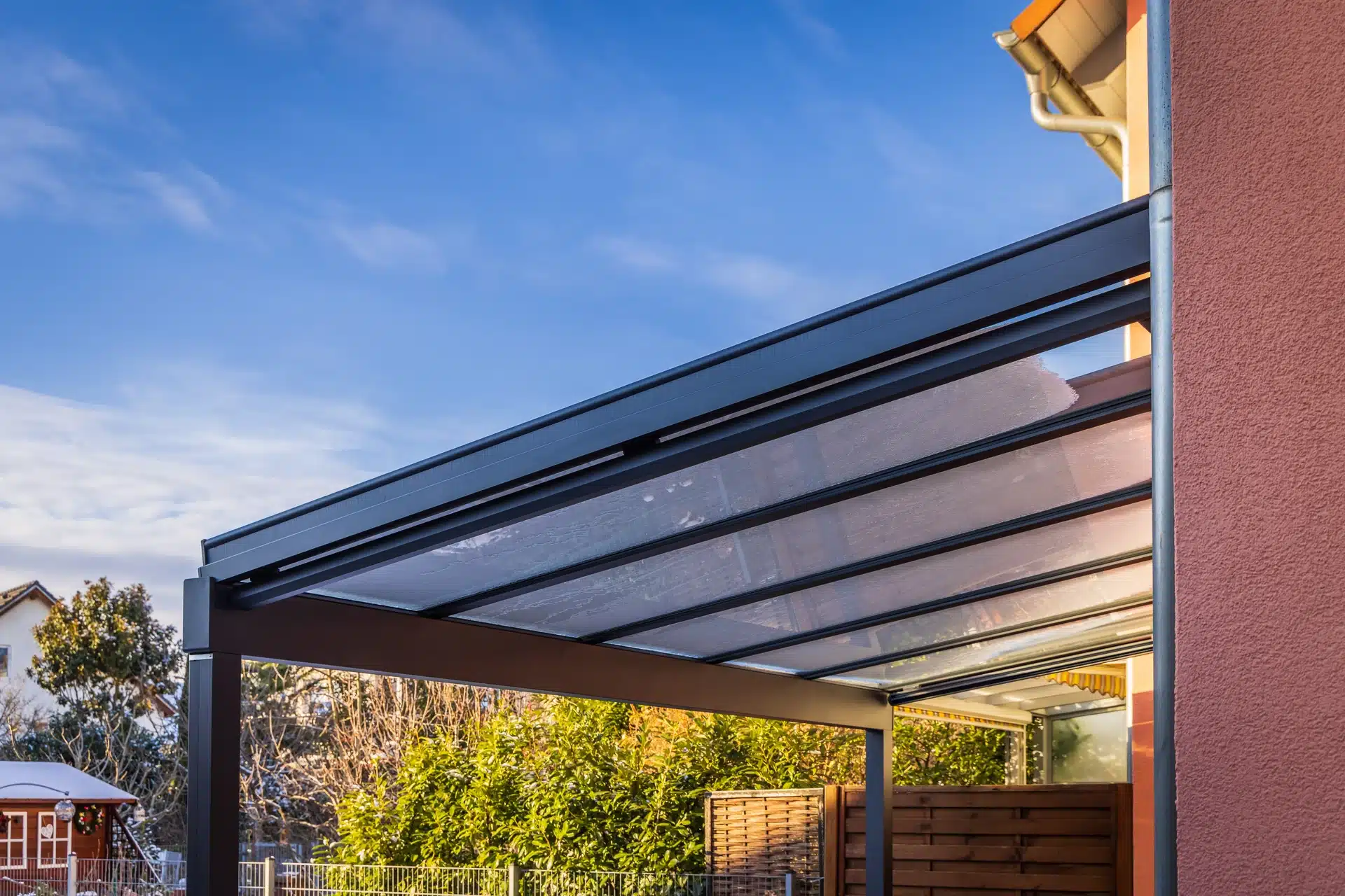Pergola moderne en aluminium noir avec toit en verre givré adossée à une maison sous un ciel bleu dégagé.