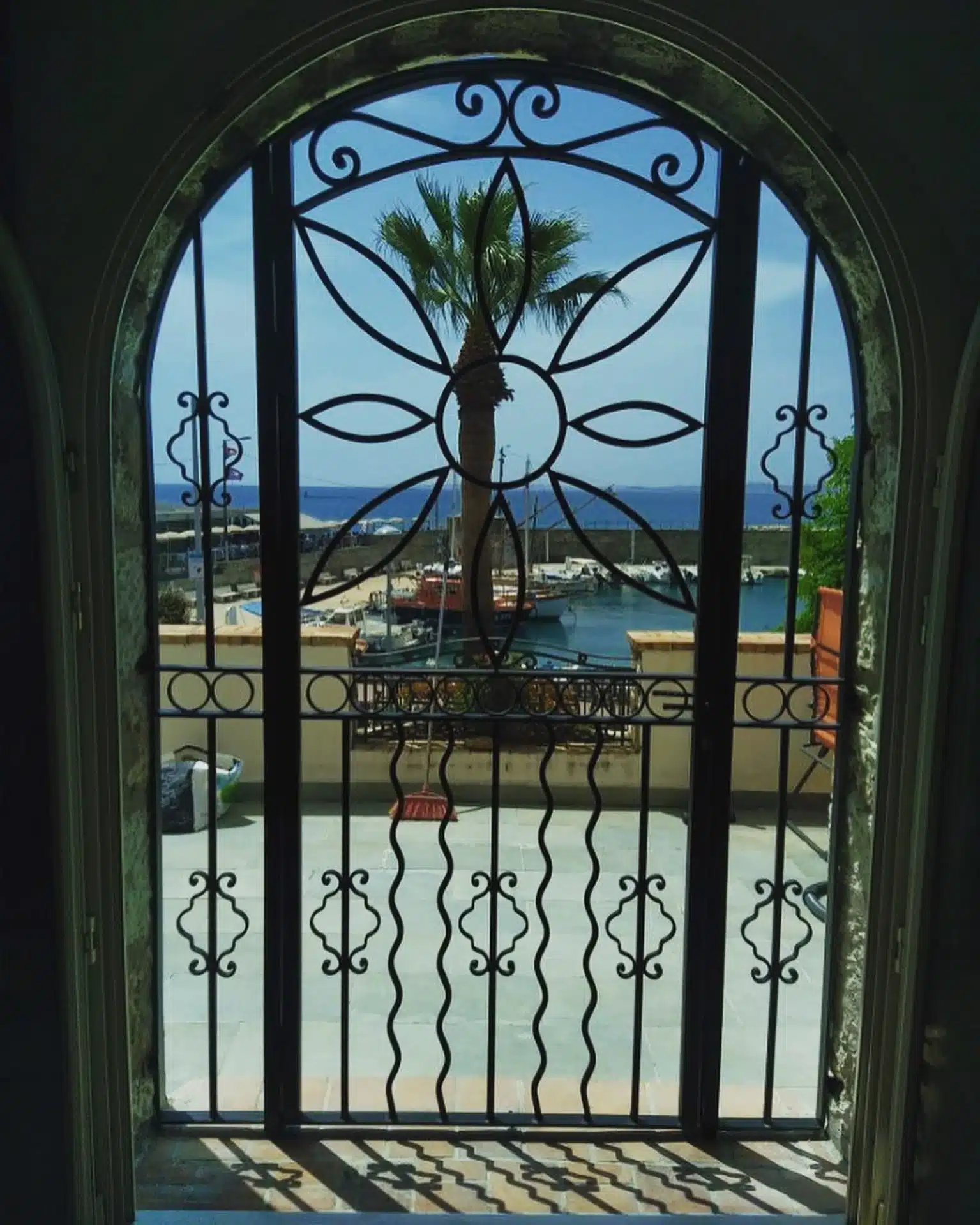 Vue d'un port méditerranéen à travers une grille en fer forgé décorative avec palmier, bateaux et mer bleue.